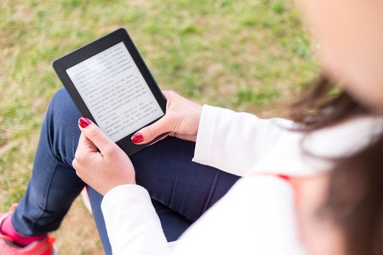 Photo of a woman sitting outside in a park reading a document on an e-book reader.