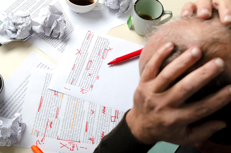 Man sitting at table holding his head in his hands while editing, highlighting, and making up paper copies of documents, with some crumpled up beside coffee cups.
