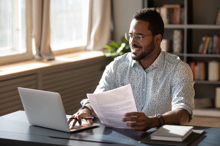 A man is holding documents, doing paperwork, preparing report or analyzing market research results, working on computer in modern workplace office.