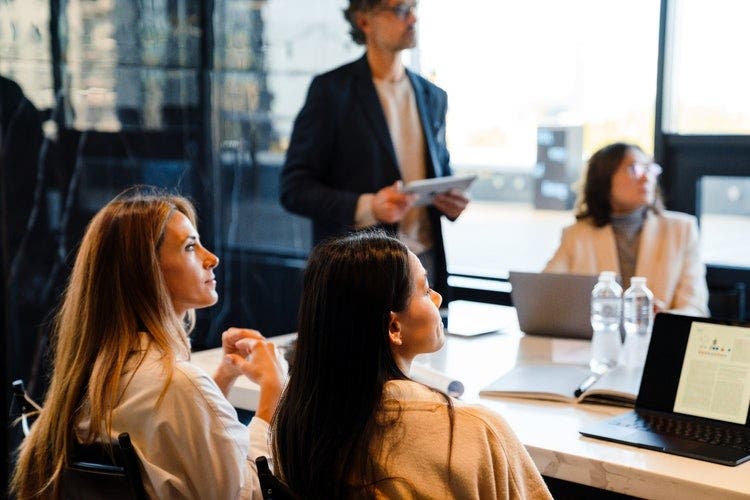 Four people attending a presentation – three women sitting at a desk with laptops, a man standing with a tablet. All are looking towards a presentation being given out of shot to the right.