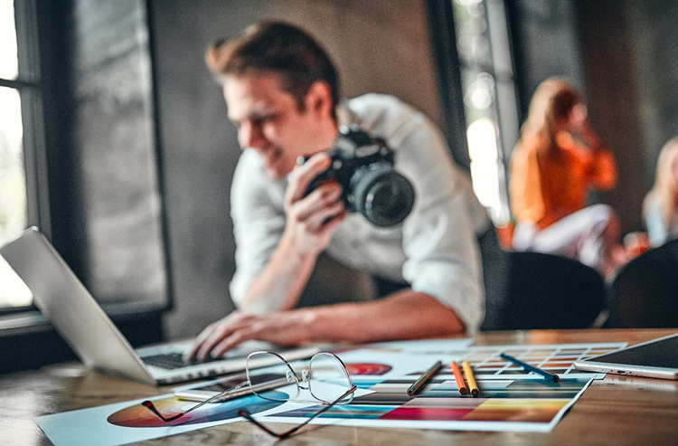 Male photographer holding a camera and working with laptop and images printed out on a desk.