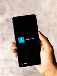 Photo of a person's hand holding a mobile phone with the Adobe Scan logo and app displayed on the screen.