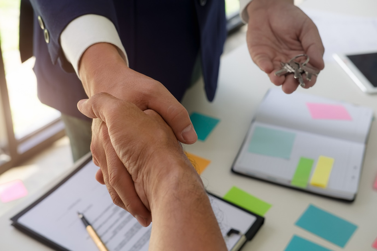 Real estate agent standing at a desk with a paper copy of a contract, hard copy diary, and sticky notes on the diary and desk. In one hand he's holding a set of keys, in the other he's shaking hands with a client.