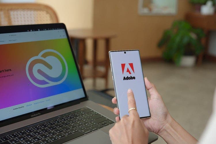 Picture of the hands of a woman holding a smartphone with the Adobe logo on the screen. On a desk in front is a laptop with the Adobe Creative Cloud homepage on the screen.