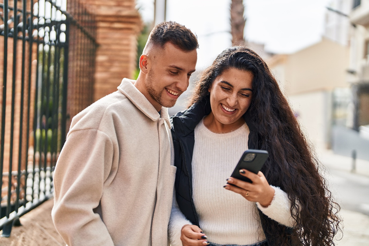 A couple smiling and looking at a smartphone while standing outside on a street.