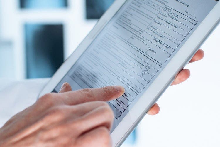 A person's hands holding a tablet with an electronic form with visible on screen. The person is completing multiple form fields on the document with one hand.