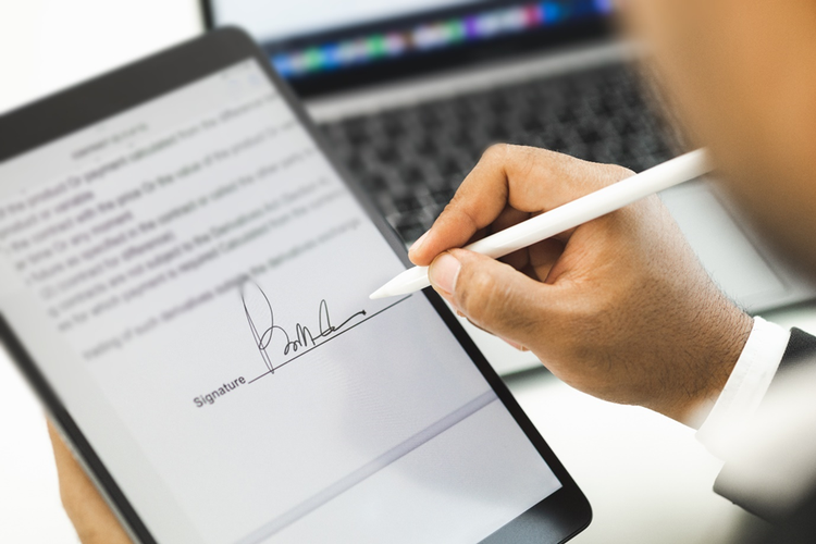 Person singing an agreement with a stylus on a tablet, with a laptop in the background.