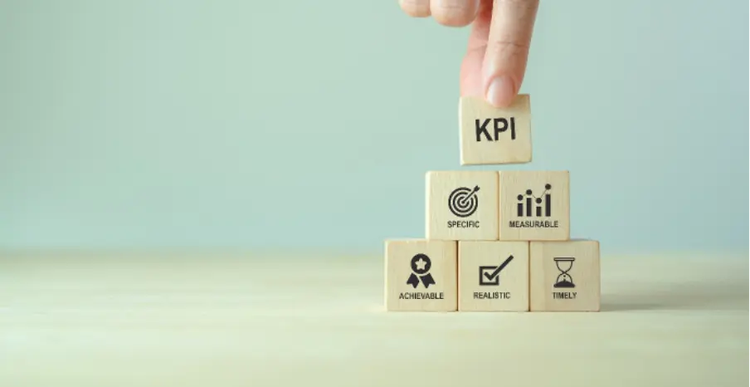 Photo of a person’s hand stacking six wooden blocks into a pyramid with KPI at the top, and blocks representing specific, measurable, achievable, realistic and timeliness underneath.