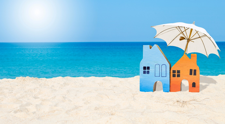 Two small wooden miniature houses on sand at the beach. One is in the sun, the other is shaded by an umbrella.