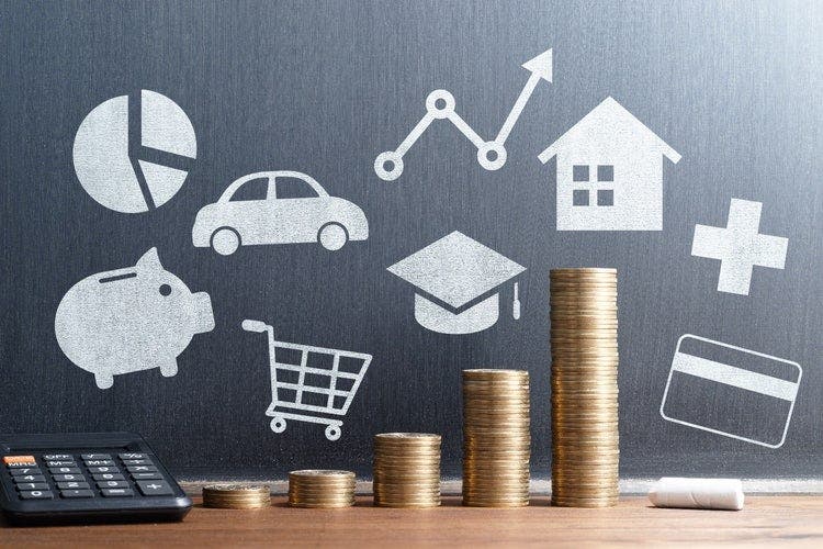 Table with a calculator, coin stacks increasing in height, and a piece of chalk. A blackboard behind the table has images of a shopping trolley, car, graduation cap, credit card, house, first aid cross, and pie chart.