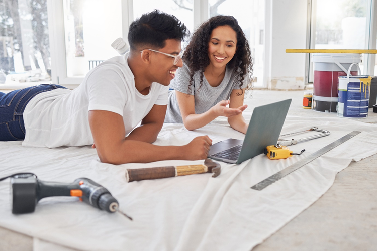 Photo of a young couple lying on the floor looking a laptop, surrounded by maintenance tools, and home renovation supplies.