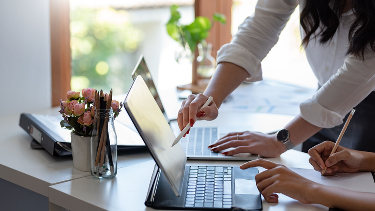 Photo showing the torso and hands of two people at a desk with laptops collaborating on the content on screen.