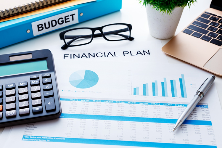 Photo of a desktop with a folder labelled budget, a calculator, laptop, financial plan, glasses, pen and potted plant.