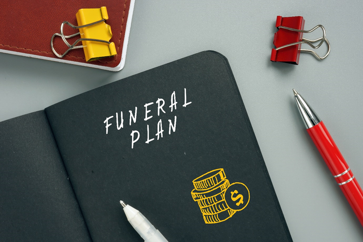 Pens and paper clips and a notebook on a desk with the words "funeral plan" and a drawing depicting money on the cover.