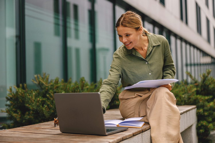 Photo of a woman sitting outside a building working on a laptop and holding paperwork.