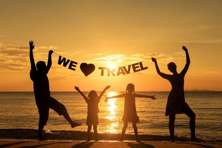 Silhouette of a family standing on a beach at sunrise or sunset. The parents are holding a string of words also in silhouette that says "We love travel".