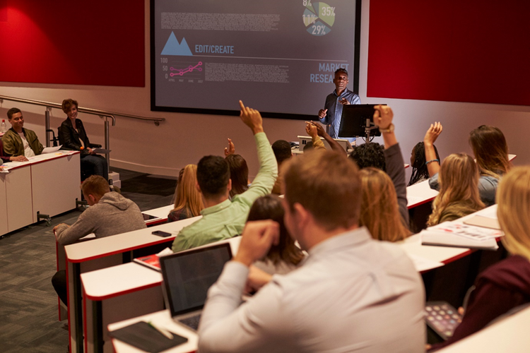 Students sitting in a lecture theatre with laptops, notebooks, and raised hands ready to ask questions.