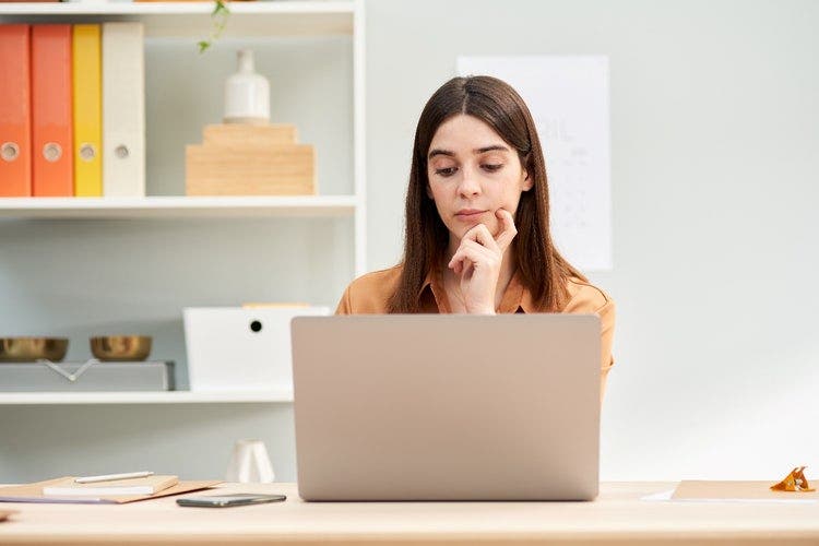 Woman sitting at a desk looking at the screen of laptop with a thoughtful expression on her face.