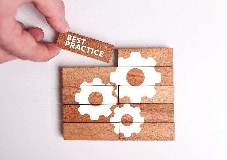 Person's hand completing a wooden puzzle with pictures of three cogs. The completing piece of the puzzle has the words "best practice".