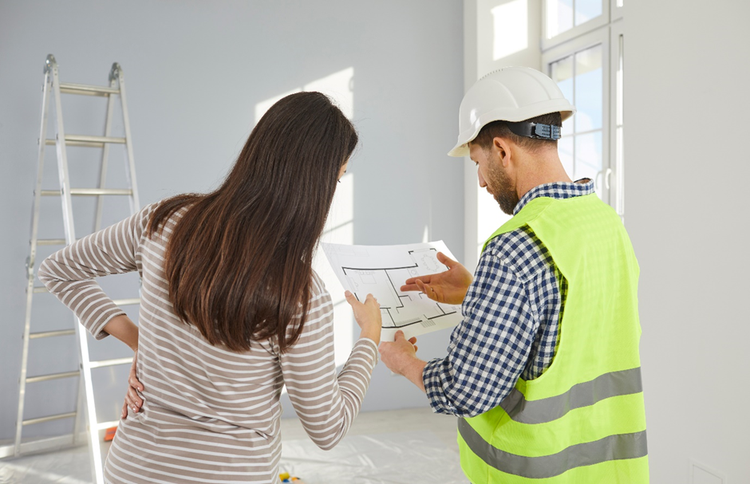 Photo of a woman inside a home reviewing house plans with a contractor in a high viz vest and hardhat.