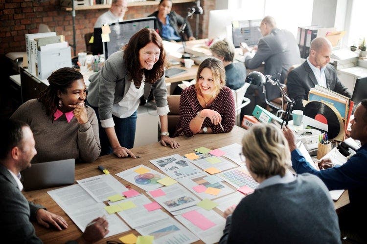 Photo of a team working in an office sitting at desks and around a table. Laid out on the table are multiple documents covered in coloured post-it notes.