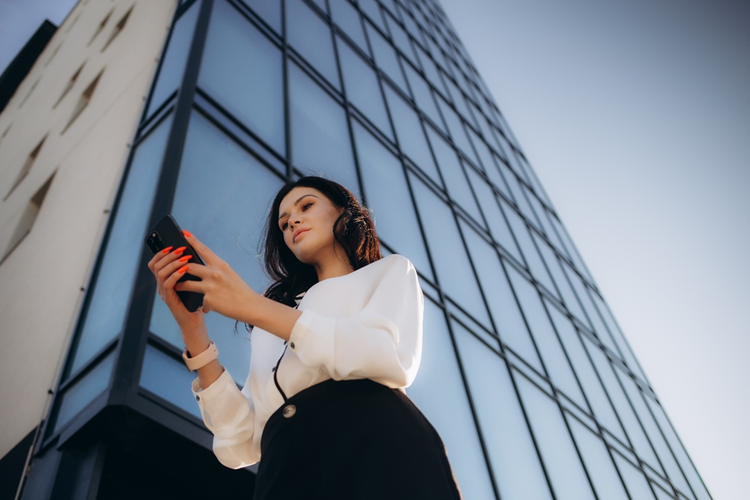 Photo of a woman standing outside a tall building looking down at a mobile phone.