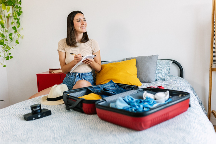 Photo of a woman sitting on bed packing her suitcase. She is smiling with a notebook in her hand for checking the holiday packing list.