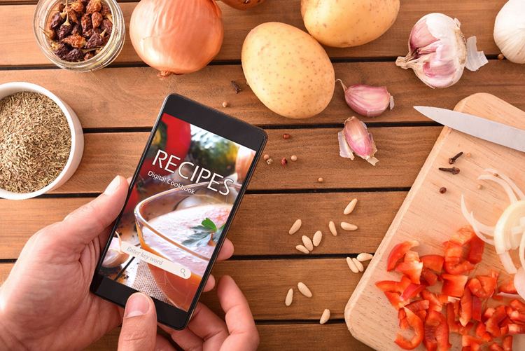 Hands of a person holding a mobile phone displaying the words "Recipes: Digital Cookbook" above a table with a chopping board and fresh ingredients.