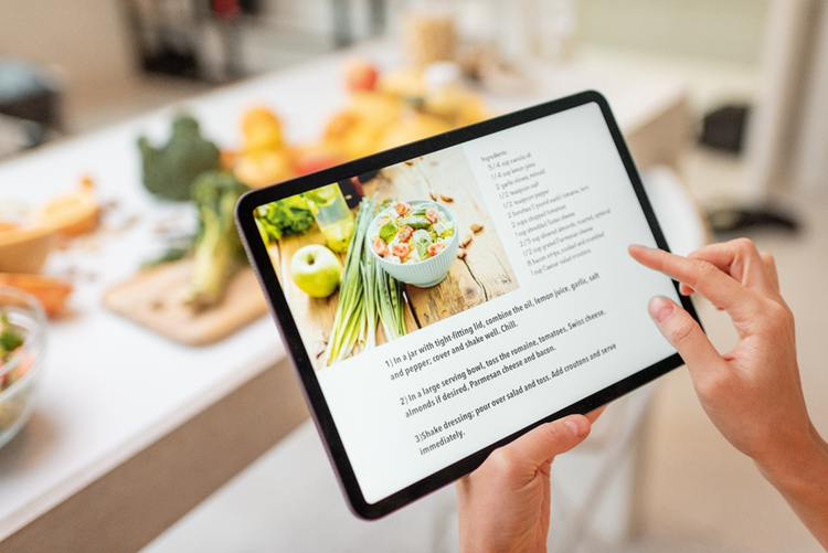 Person in a kitchen holding a table with a recipe displayed on screen and fresh vegetables on the benchtop.