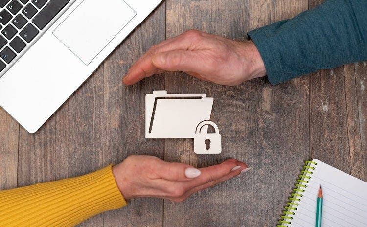 Two hands are cradling an image of a shared folder with a padlock. A male's hand is on top, a females on the bottom. In the corners of the desk are a laptop and a notepad.