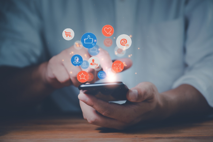 Hands of a person holding a mobile phone with conceptual icons floating above the screen showing likes, messages, shopping cart, locations and notifications.