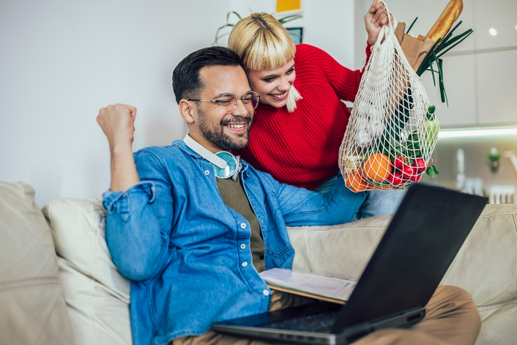 Smiling Couple sitting at home on the couch looking at a laptop and holding a string bag with fresh groceries.