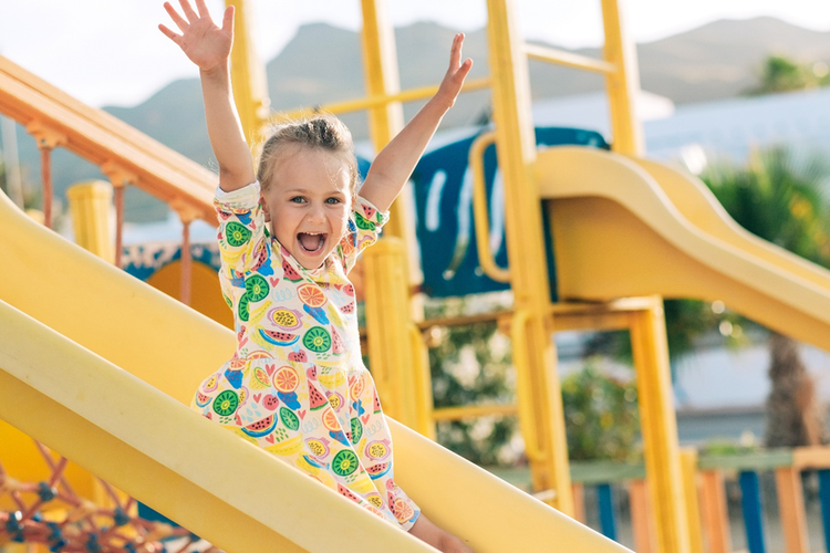 Photo of a young girl on a slide in a playground with her hands in the air and smiling.