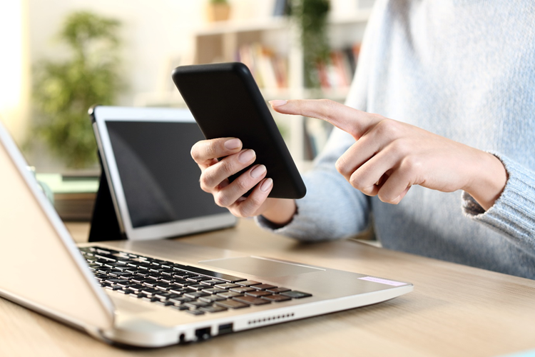 Photo of a woman’s torso and hands sitting at a desk with multiple devices including a smartphone, laptop and tablet.