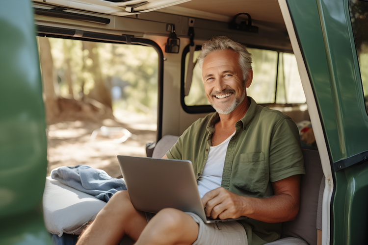 Photo of a man sitting in the back of a camper van with laptop and trees in the background.