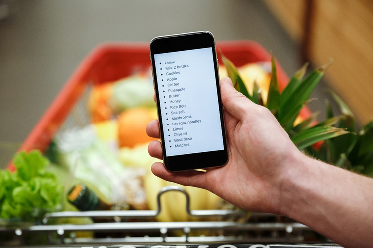 Photo of a person in a supermarket with shopping trolling holding a mobile phone with a grocery list displayed on the screen.