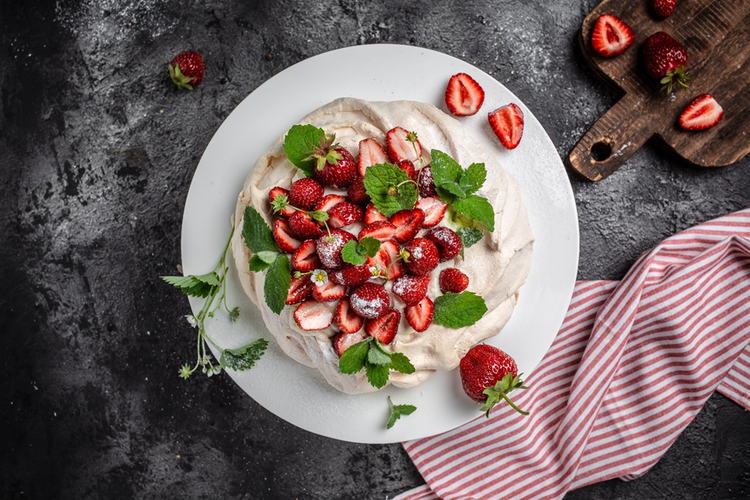 Photo of a pavlova topped with strawberries and mint beside a wooden chopping board and red and white striped tea towel.