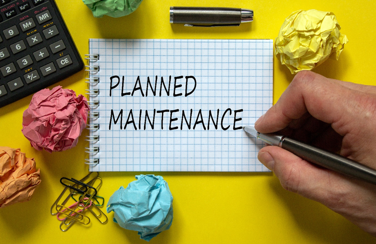 Person's hand holding a pen and writing "Planned Maintenance" on a notepad beside a calculator and crumped up pieces of paper.