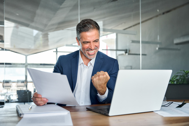 Man sitting at a desk with a laptop and paperwork, smiling at the computer screen and making a hand gesture indicating success/good results.