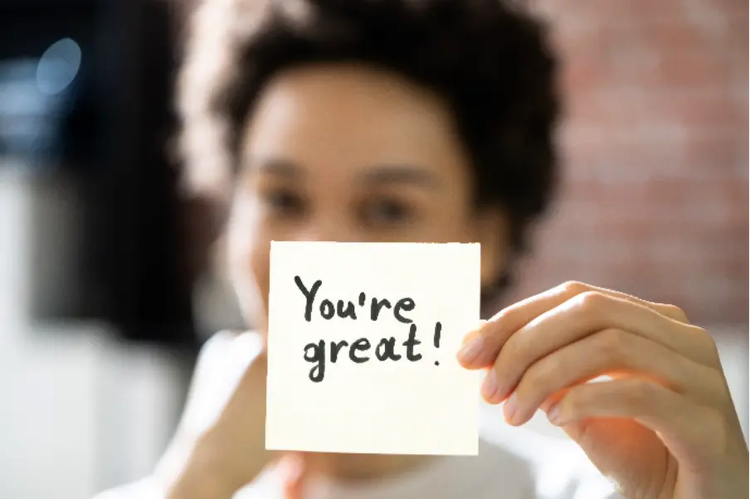 Photo of a woman holding a sticky note with the words “You’re great” written on it.