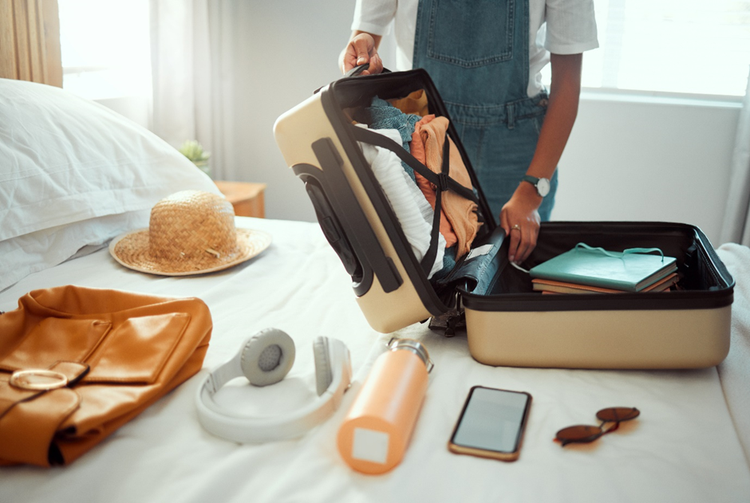 Photo of a woman standing in bedroom and packing a suitcase on a bed with holiday items such as a sunhat, day pack, clothing, sunglasses, mobile phone and refillable water bottler.