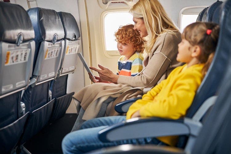 Woman and two children sitting in a plane. The woman is showing what's on the screen of a tablet to the children.