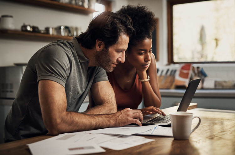 Photo of a couple sitting in a kitchen looking at the screen of a laptop with copies of receipts on the table.