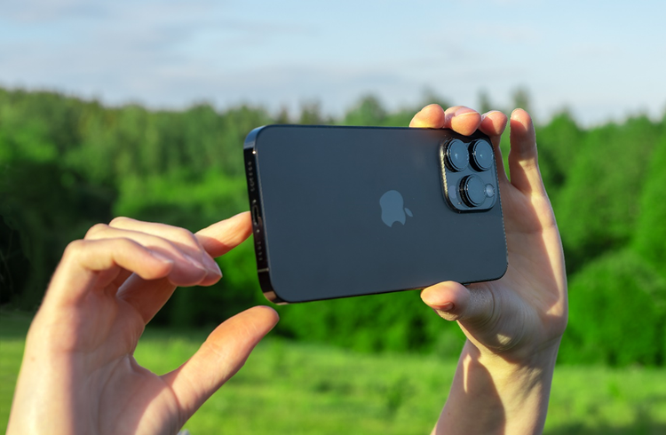 A person uses a black iPhone to take photos in an open field surrounded by lush greenery.