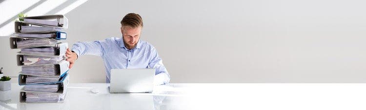 Male sitting at desk with laptop and pushing aside a stack of folders containing paperwork.