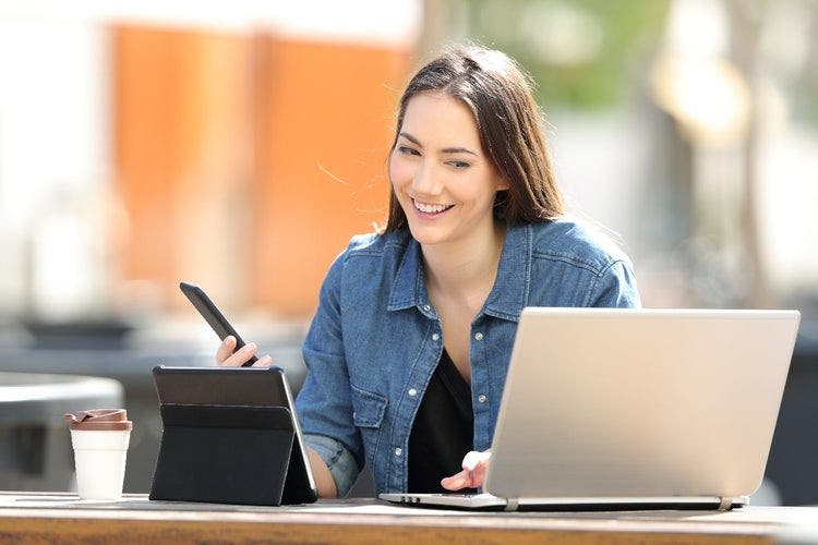 Woman sitting at a desk and smiling while using multiple devices - a phone, laptop and tablet.