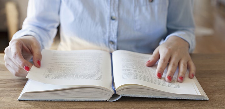 Person in a light blue shirt sitting at a wooden table reading a book.