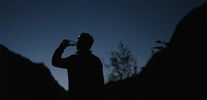 A silhouette photo of a person drinking a bottle of water at nighttime with mountains in the background