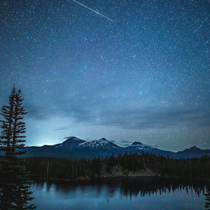 The Milky Way lighting up the night sky for lake in the middle of a forest with snowy mountains in the background