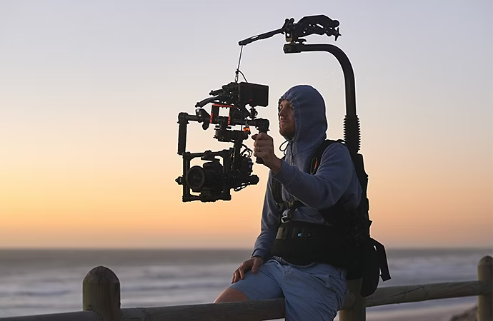 Um videógrafo sentado em uma cerca com um equipamento steadicam preso a ele